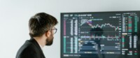Man Observing Stock Market Data On A Screen And Holding A Tablet, Representing Financial Analysis And Trading.